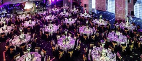 Un vaste espace intérieur aménagé pour les événements, avec de nombreuses tables rondes, chacune recouverte d’une nappe blanche et ornée d’un ruban doré, le tout éclairé d’une lumière violette.