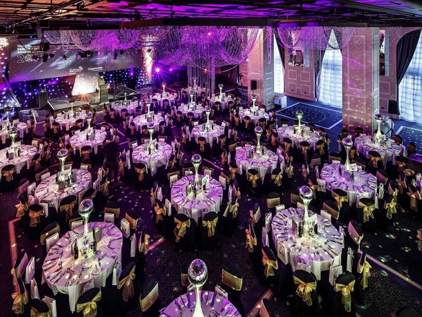 A large indoor event space with multiple round tables, each set with white tablecloths and gold sashes, under purple lighting. - Mercure Manchester Piccadilly Hotel (Manchester)