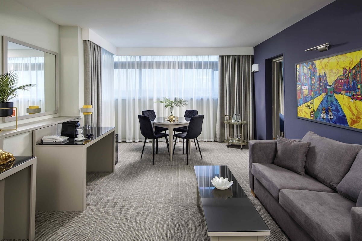 A hotel room with a dining area, a sofa, a coffee table, and a painting on the wall.