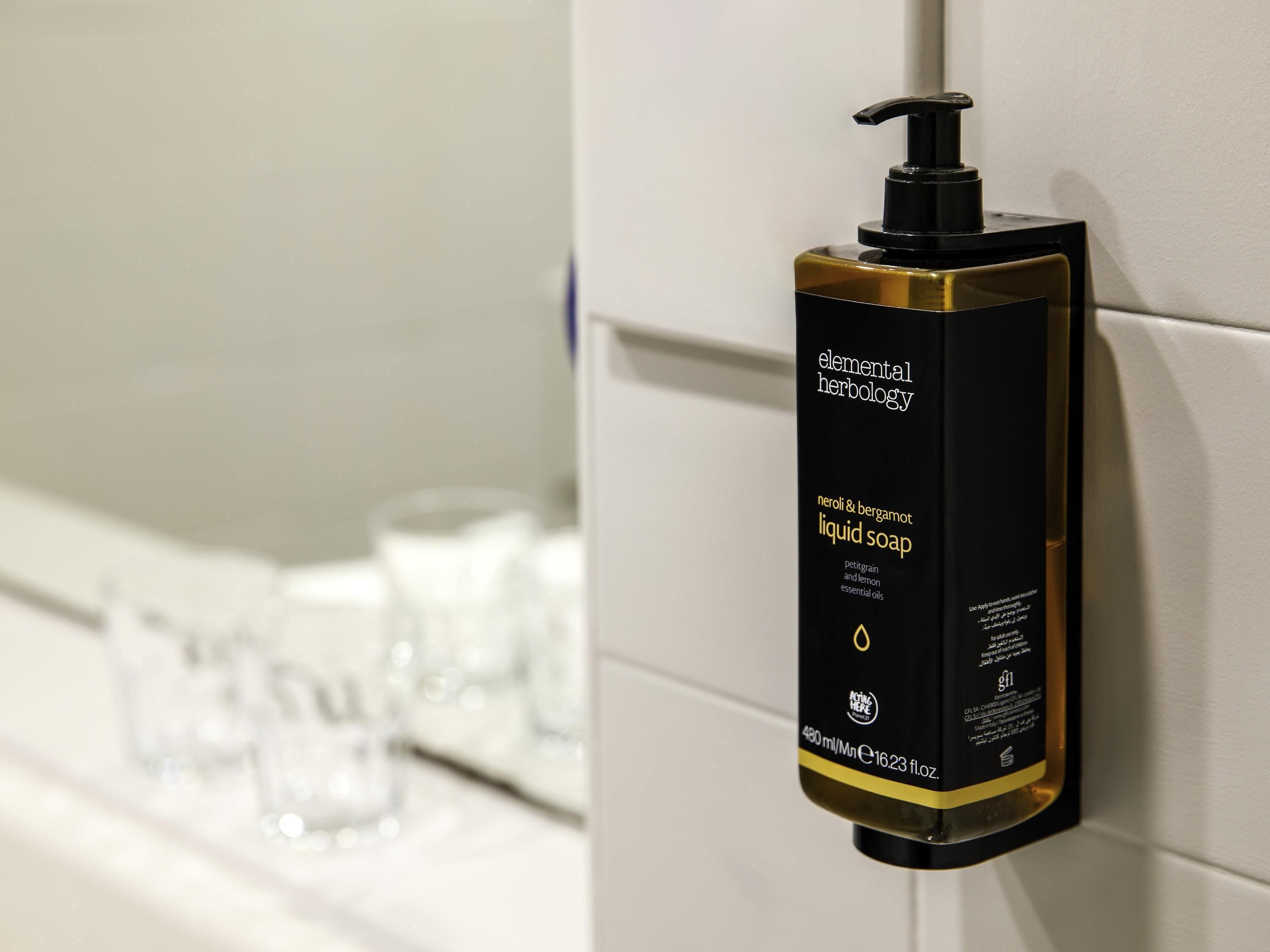 A black and gold bottle of liquid soap with a pump dispenser, labeled 'elemental herbalology,' mounted on a white tiled wall.