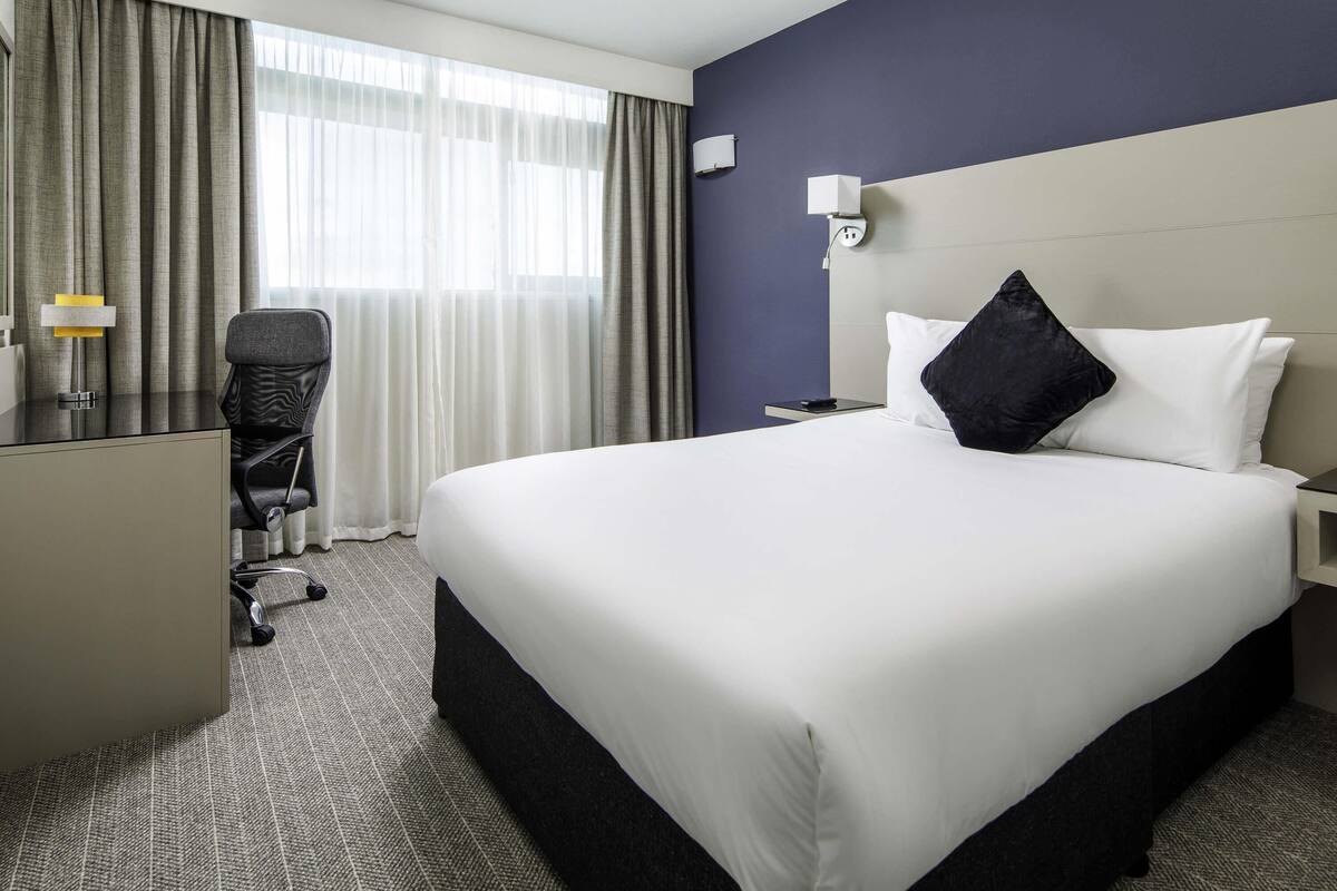 Een hotelkamer met een groot bed, een bureau, een stoel, een nachtkastje en een raam met gordijnen.