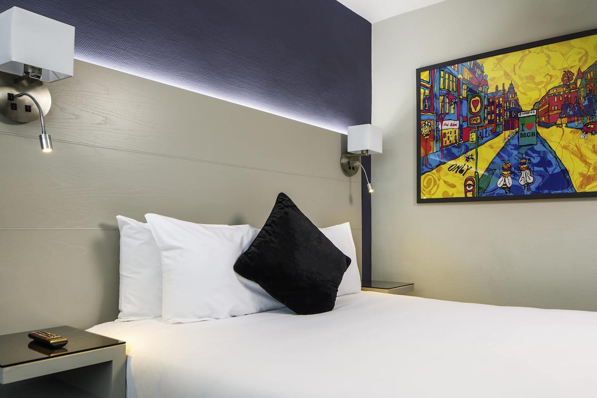Une chambre d’hôtel avec un lit, des oreillers blancs et noirs, une table de chevet avec une télécommande, une lampe fixée au mur et un tableau coloré.