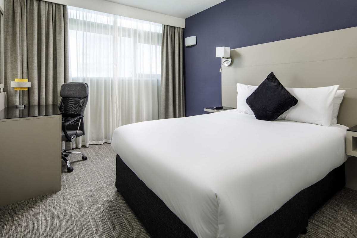 Une chambre d’hôtel avec un grand lit, un bureau, une chaise, une table de chevet et une fenêtre avec des rideaux.