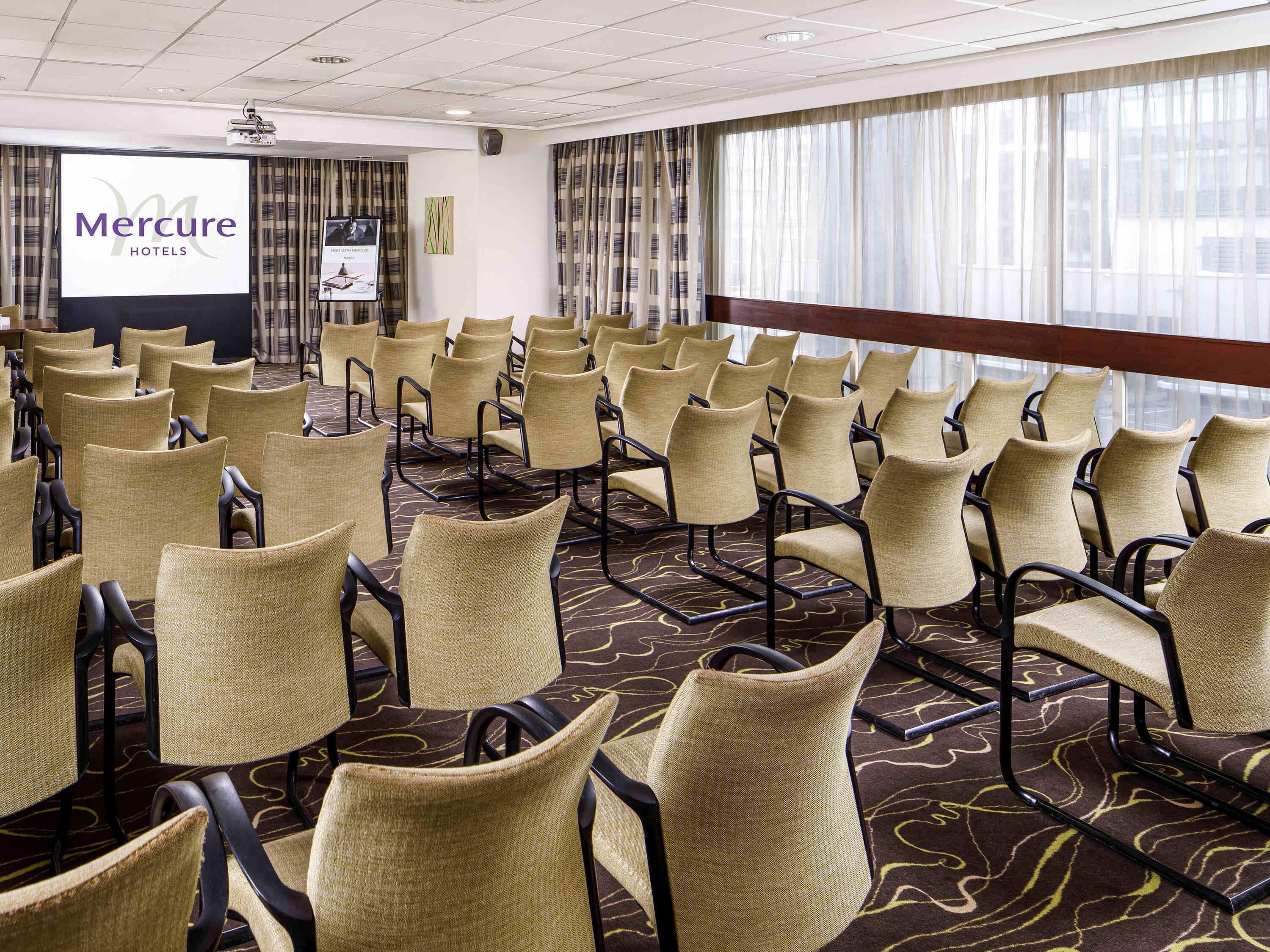 Een vergaderzaal met rijen stoelen, een scherm waarop 'Mercure Hotels' staat geprojecteerd en grote ramen met gordijnen.