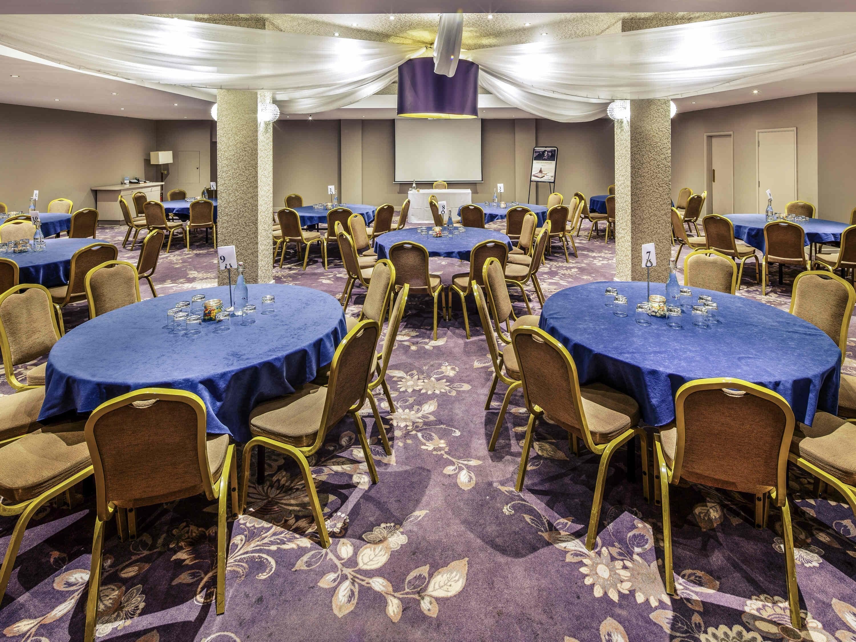 Un salón de conferencias con mesas redondas, sillas y una pantalla para proyector.