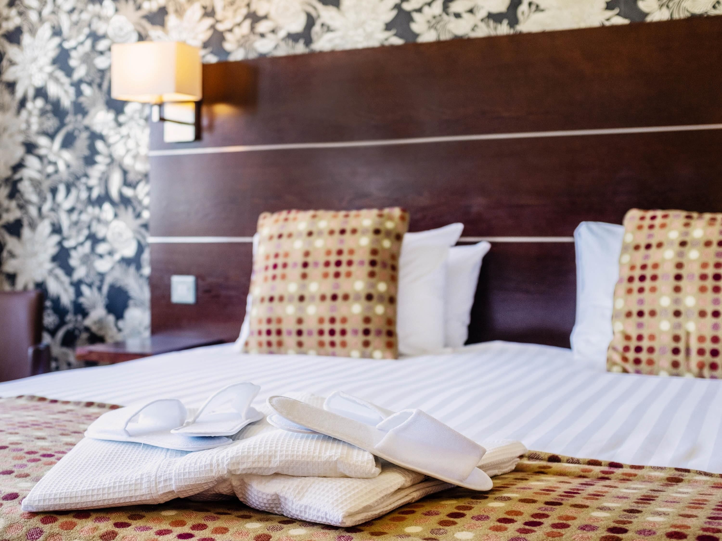 A neatly made bed with pillows, a pair of slippers, and a bedside lamp.