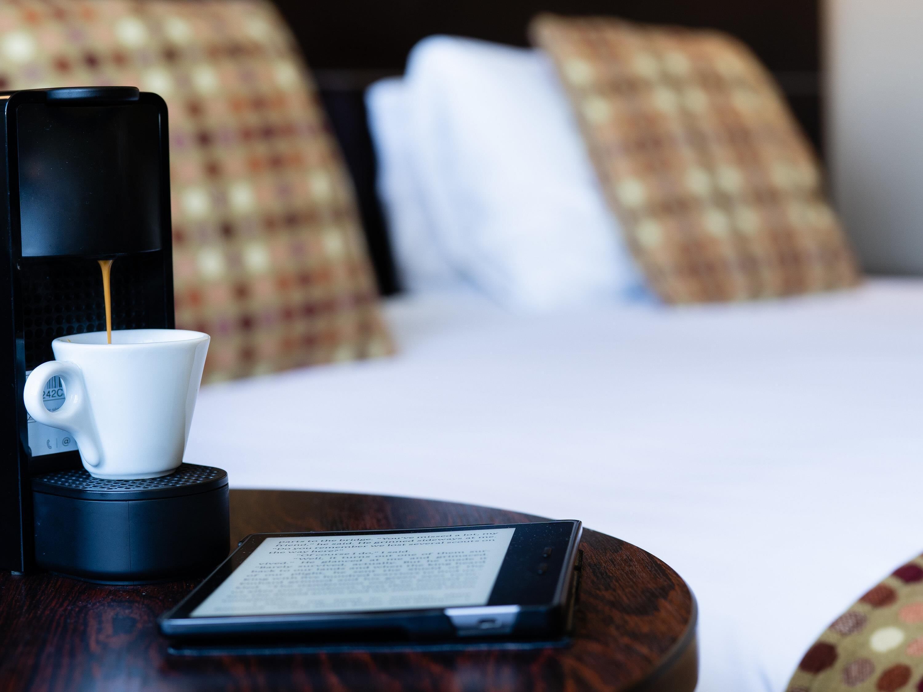 A coffee maker with a cup of coffee, a tablet, and a bed with pillows in the background.