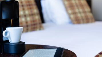 A coffee maker with a cup of coffee, a tablet, and a bed with pillows in the background.