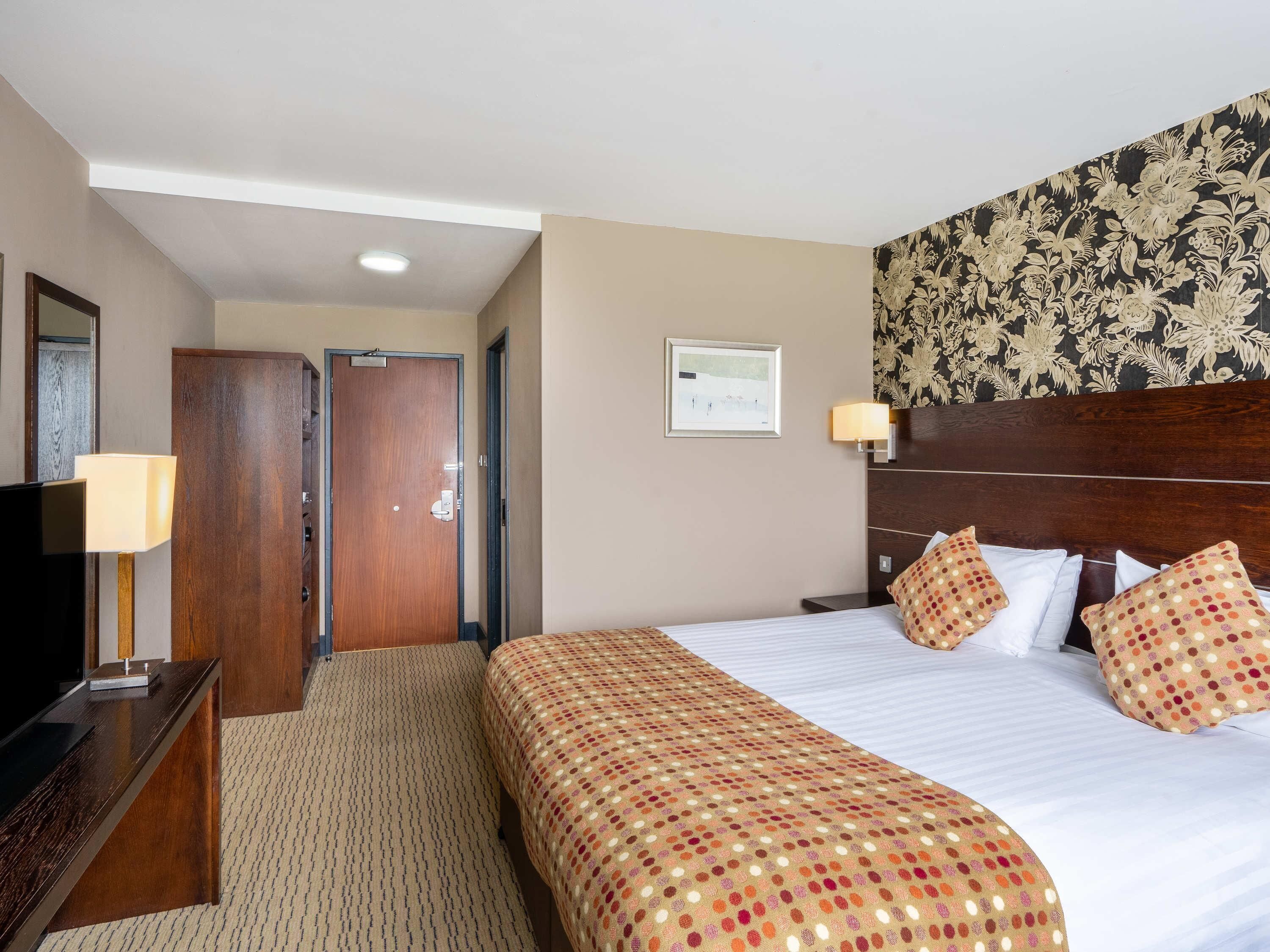 A hotel room with a bed, a desk with a lamp, a wooden wardrobe, a door, and a wall with a framed picture.
