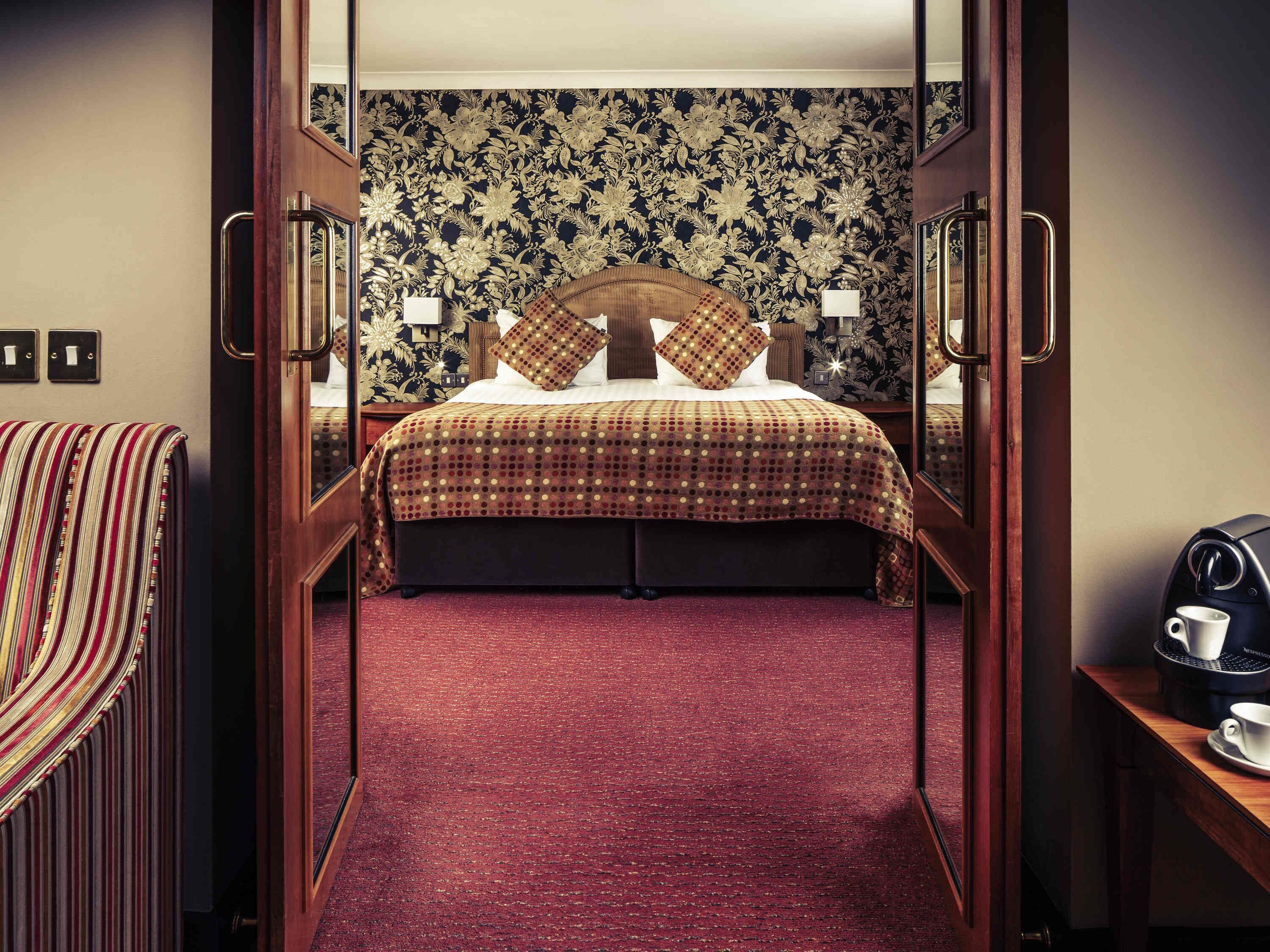 Una camera d'albergo con un letto, una moquette rossa, una testiera in legno, un comodino, una lampada e una macchina del caffè.