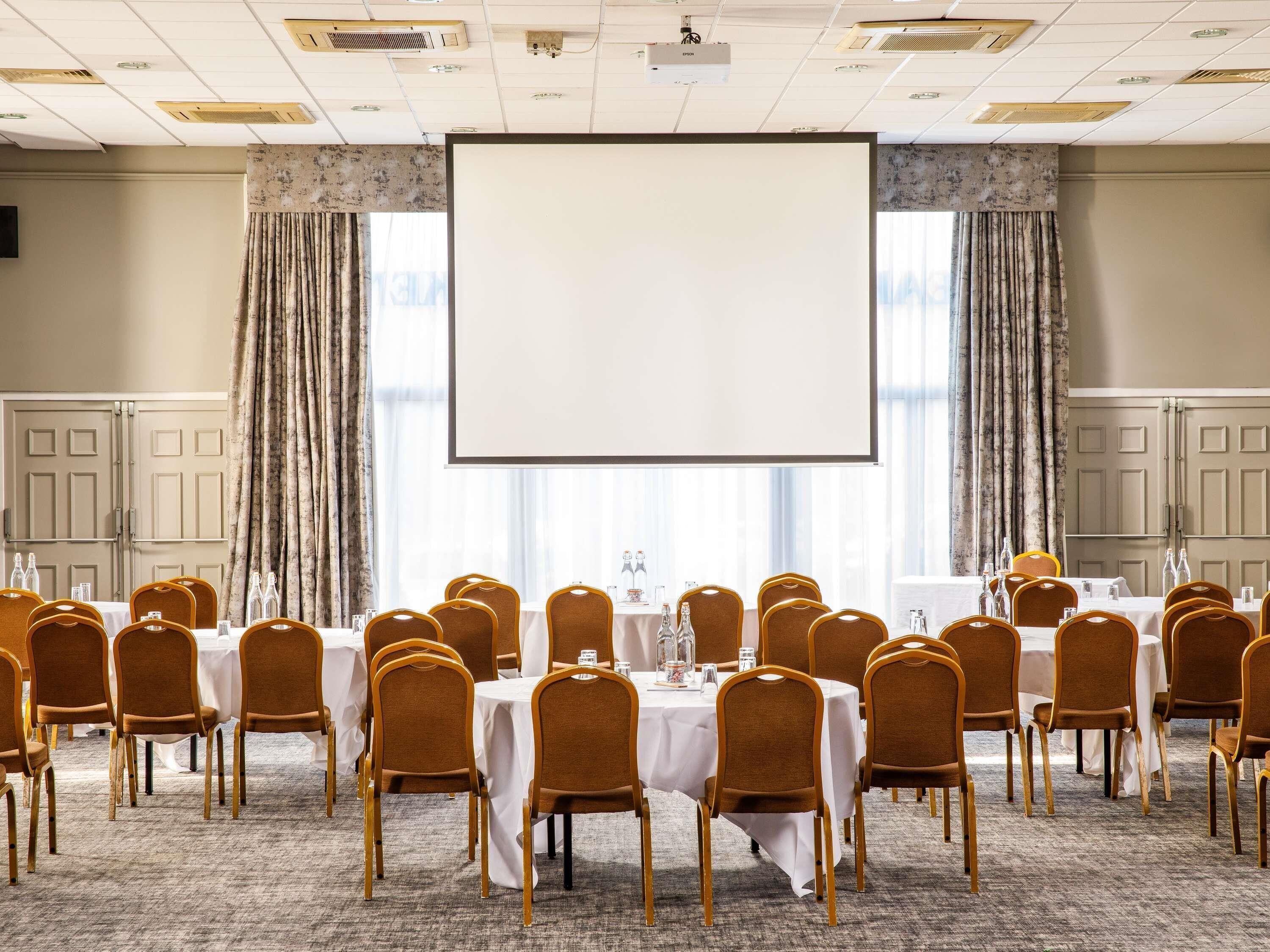 A conference room with multiple tables and chairs, a large projection screen, and curtains.