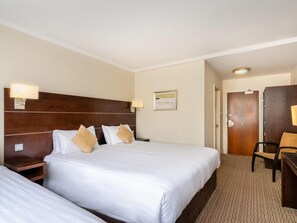 Habitación de hotel con dos camas, cabecero de madera, lámparas de noche, un cuadro en la pared y un escritorio con una silla.