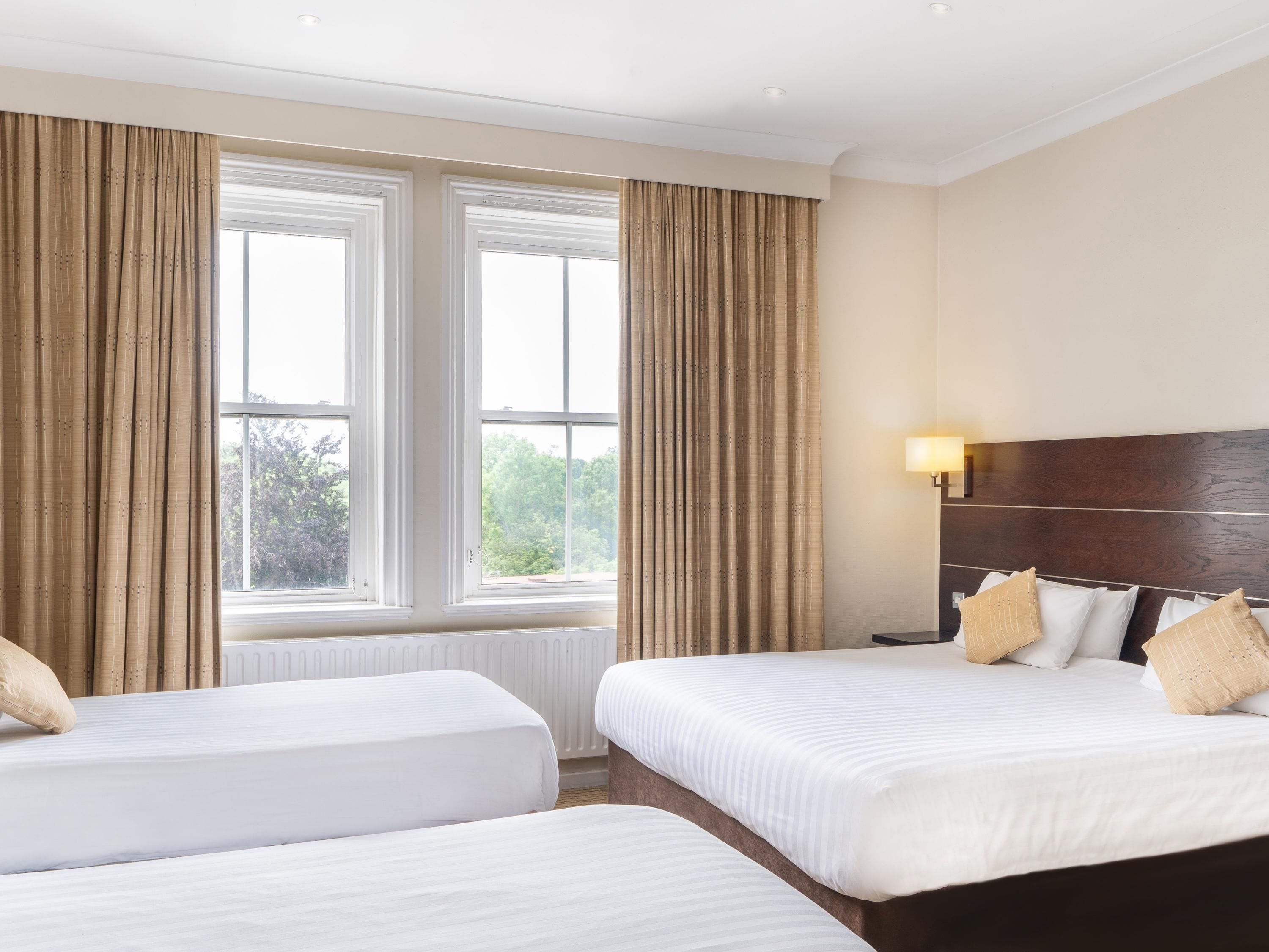 A hotel room with two beds, a large window with curtains, a nightstand with a lamp, and a view of trees outside.