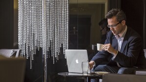 A man in a suit sitting at a table with a laptop and a glass of water, in a room with a chandelier. - The Queen's Gate Hotel (London)