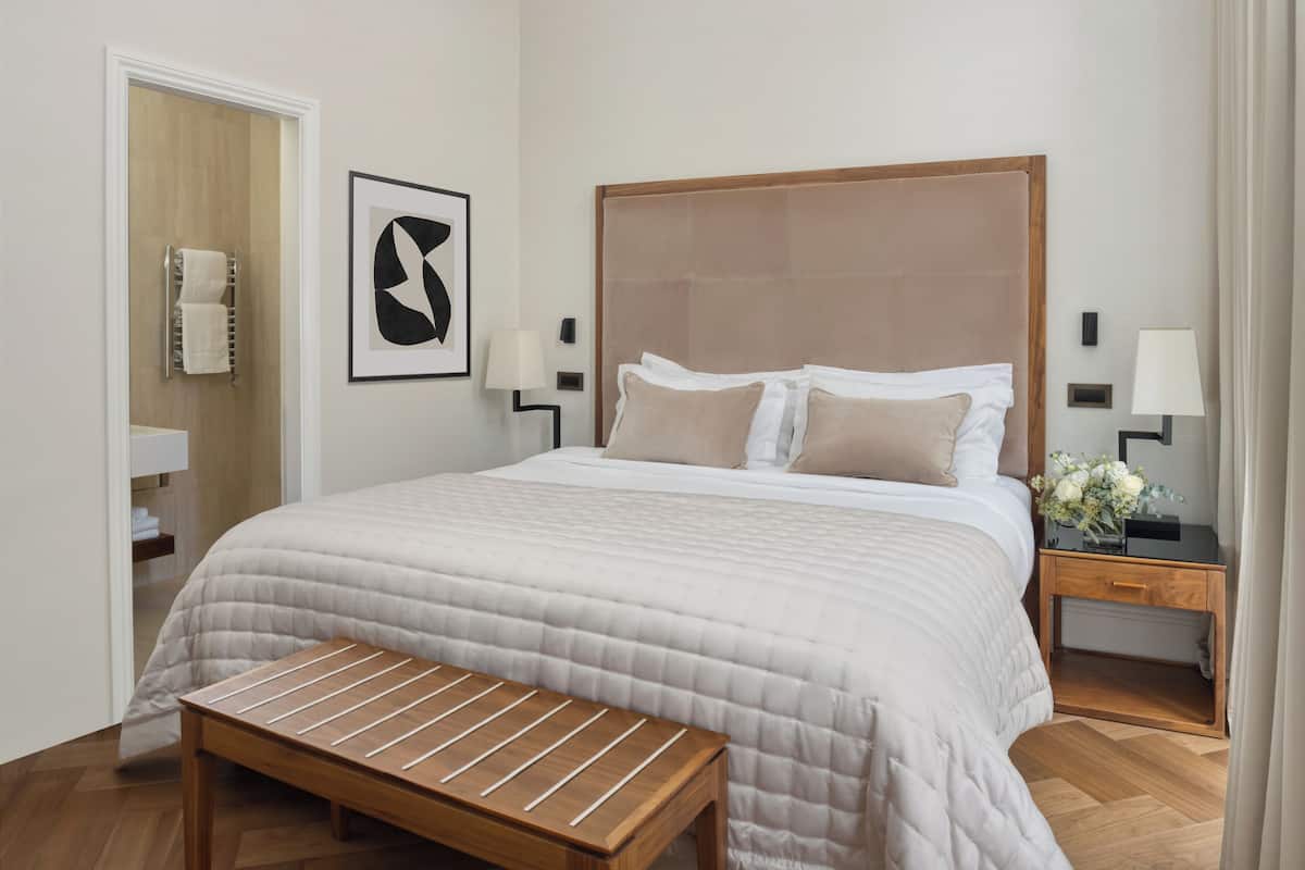 Habitación de hotel ordenada con una cama grande, un banco de madera, una mesita de noche con lámpara y una obra de arte abstracta enmarcada en la pared.