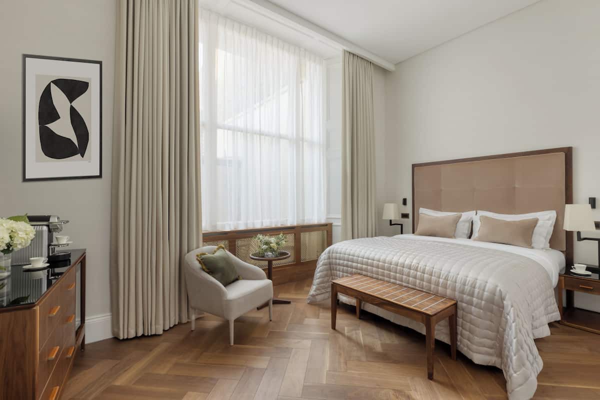 Habitación de hotel moderna con una cama grande, una zona de estar con una silla y una mesita, y un ventanal con cortinas.