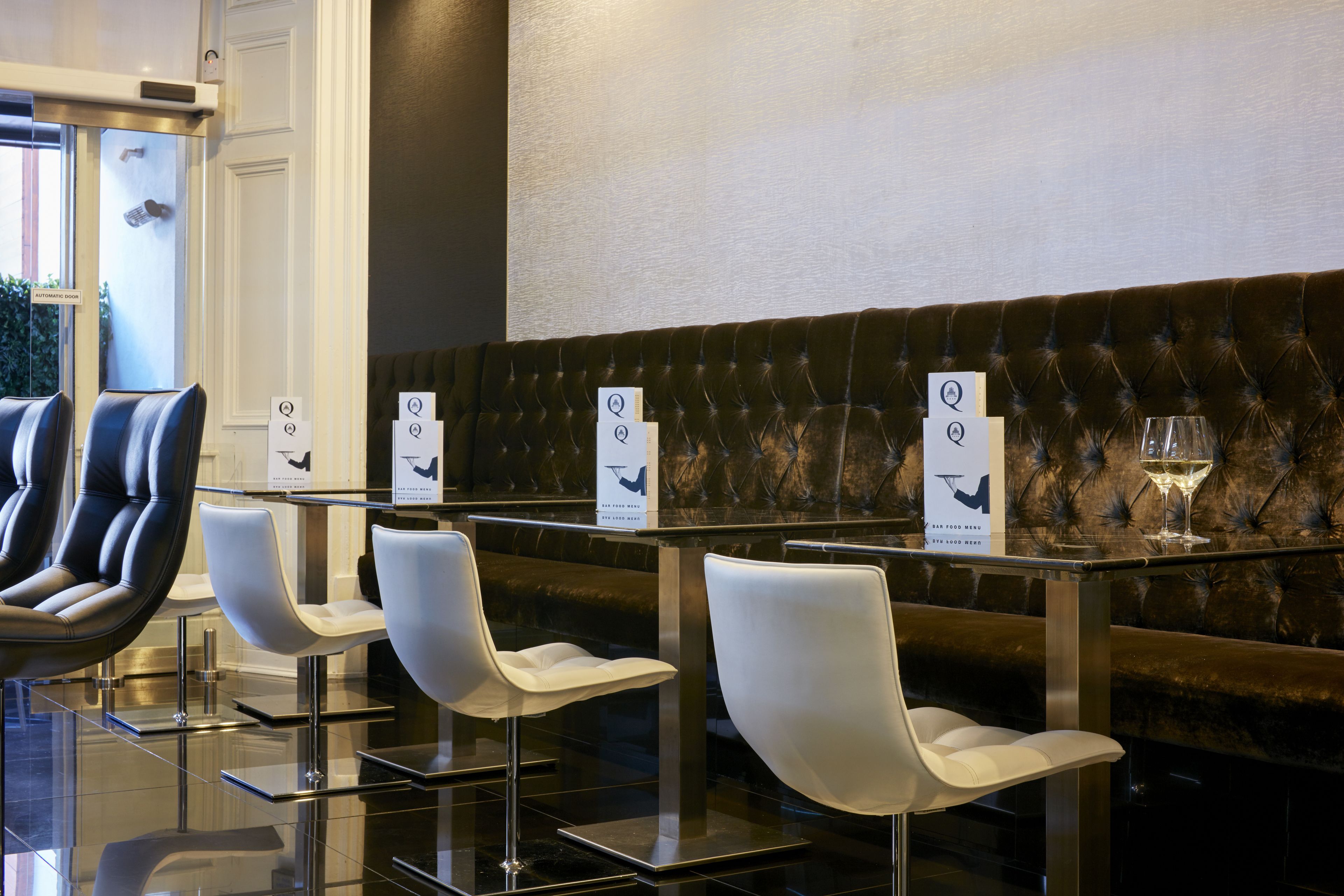 Un moderno hall de hotel con zona de estar que incluye sillas blancas y un banco tapizado oscuro.