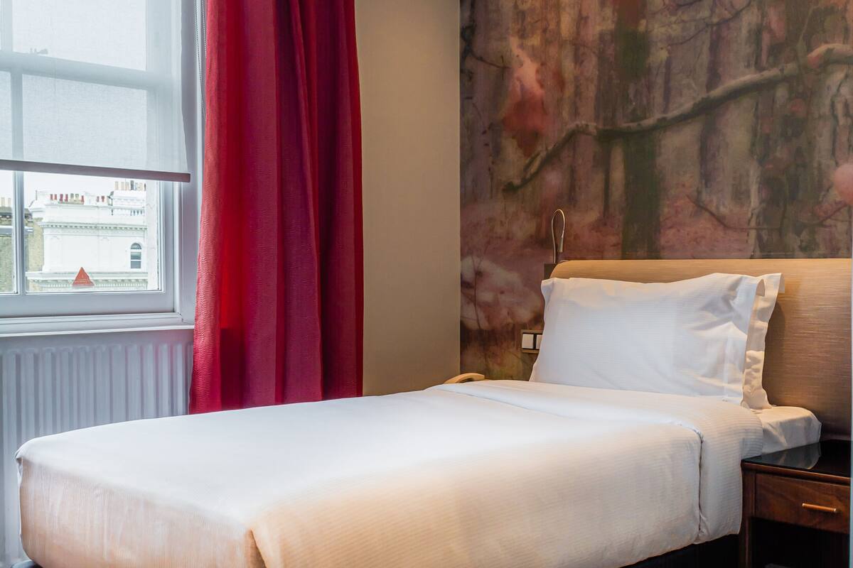 Habitación de hotel con una cama, una mesita de noche, un ventanal con cortinas rojas y un mural texturizado en la pared.