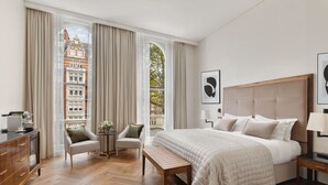 A hotel room with a large bed, a desk, a chair, a small table, and a view of buildings through large windows. - The Queen's Gate Hotel (London)