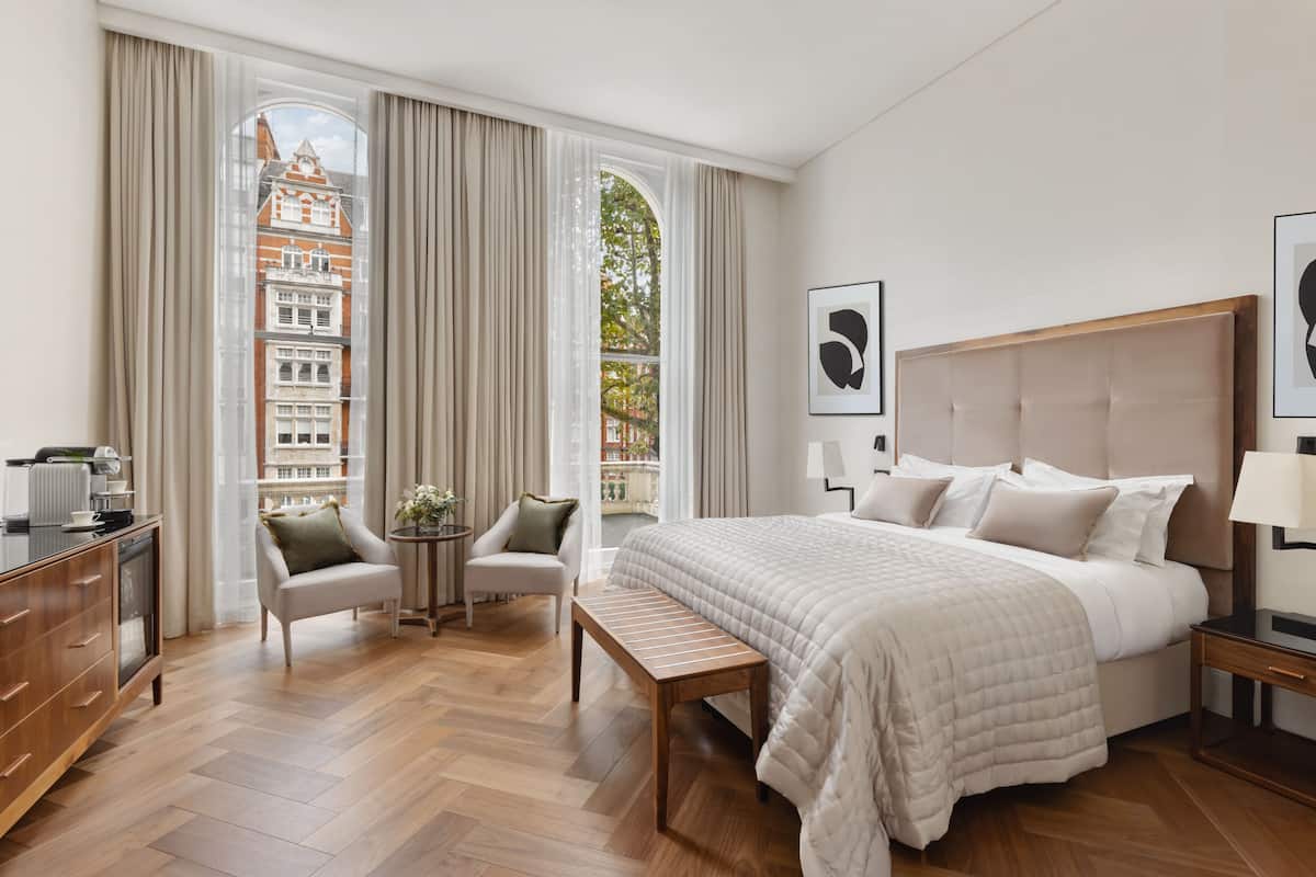 Habitación de hotel con una cama grande, un escritorio, una silla, una mesita y vistas a edificios a través de amplias ventanas.