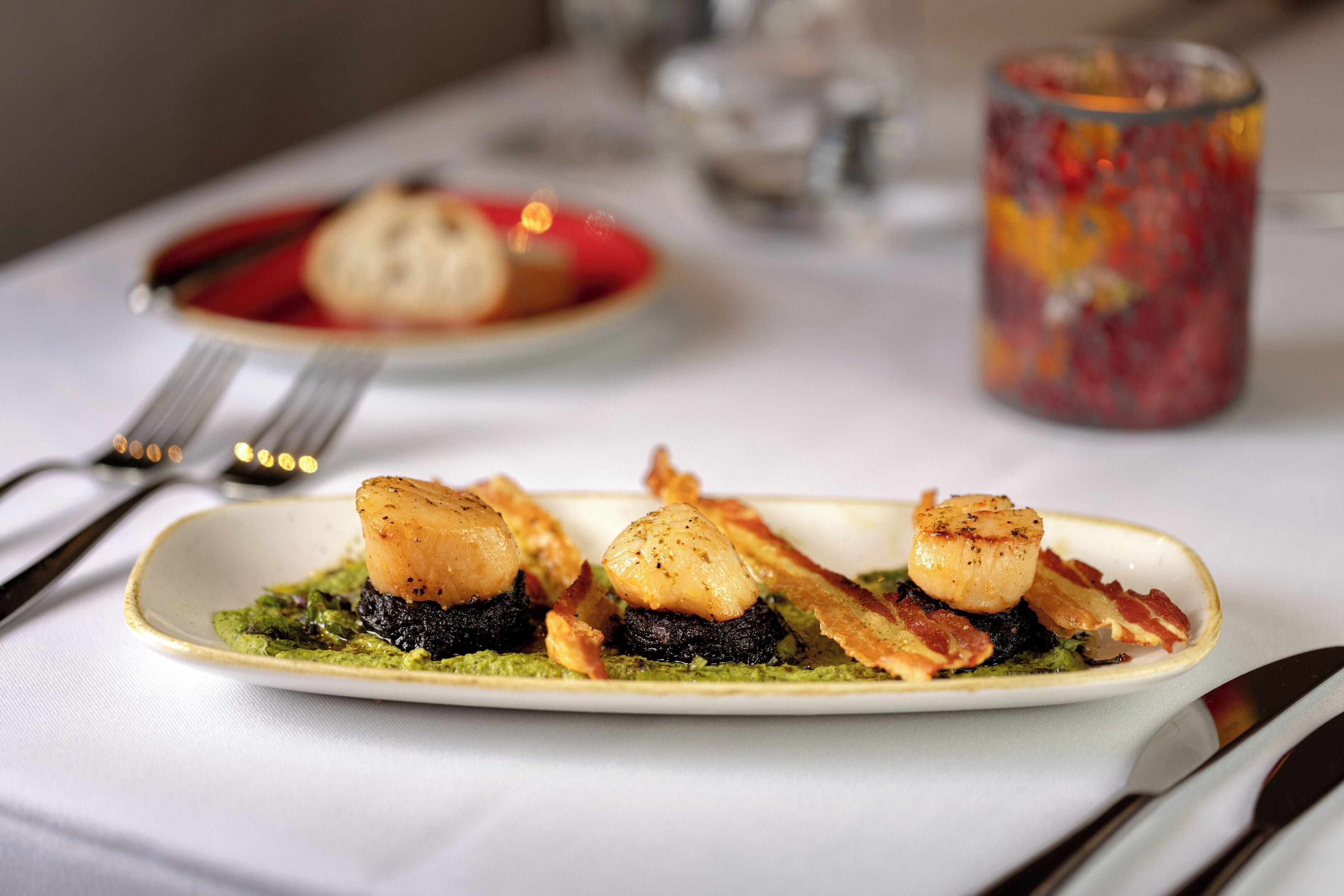 A plate of food with seared scallops and bacon on a bed of green sauce, accompanied by a glass of red beverage.