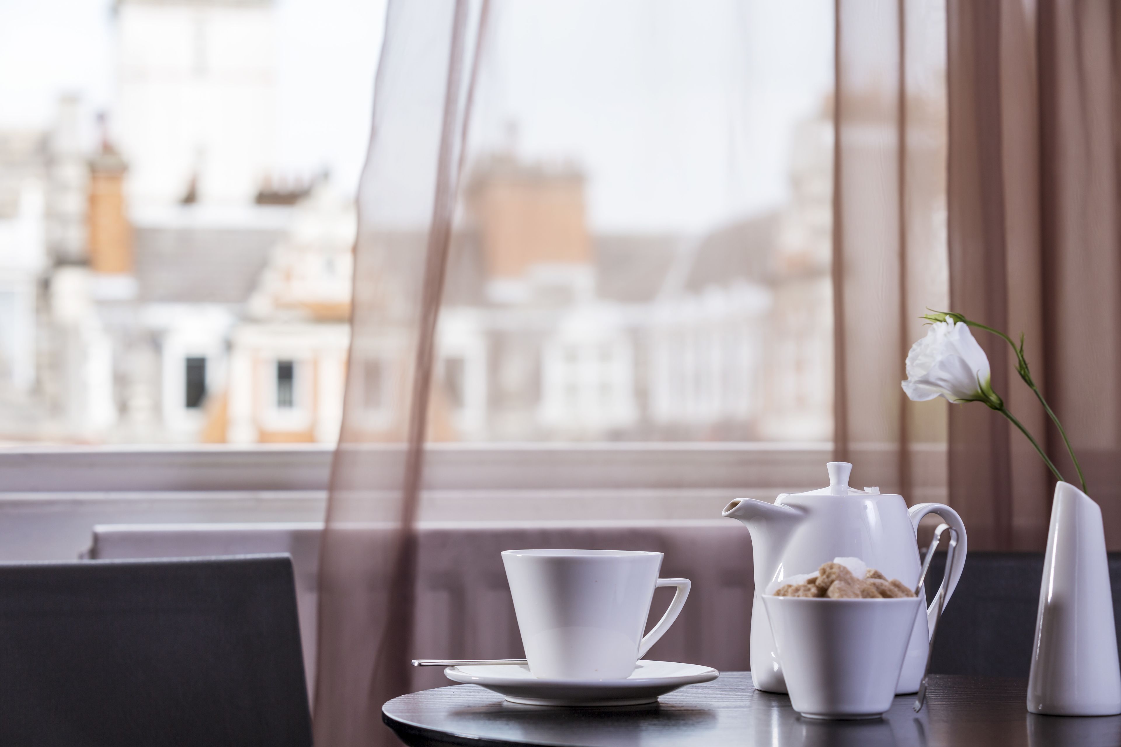 Una mesa con una taza blanca, un platillo, una tetera y un azucarero, un florero blanco con una sola flor blanca, y vistas de edificios a través de una ventana.