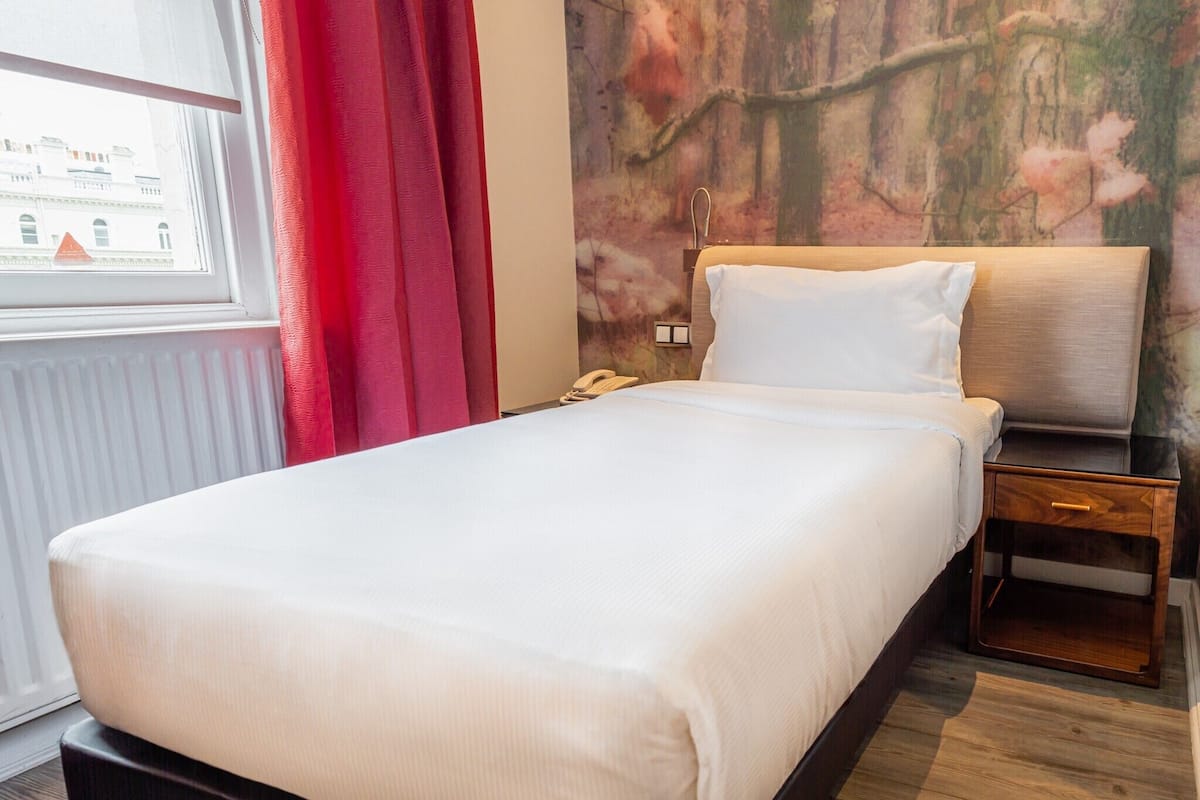 Una habitación de hotel con una cama, una mesita de noche, un teléfono y una ventana con cortinas rojas.