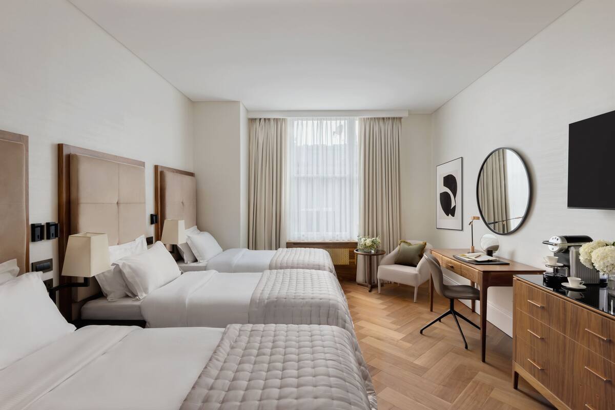 Habitación de hotel con tres camas, un escritorio, una silla y una pequeña mesa con lámpara.