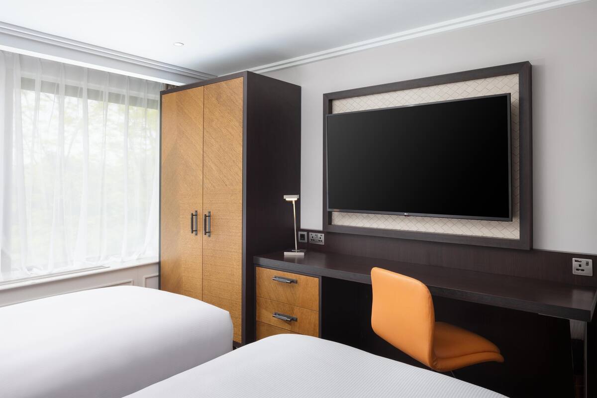 Una habitación de hotel con una cama, un escritorio, una silla naranja, un armario y un televisor en la pared.