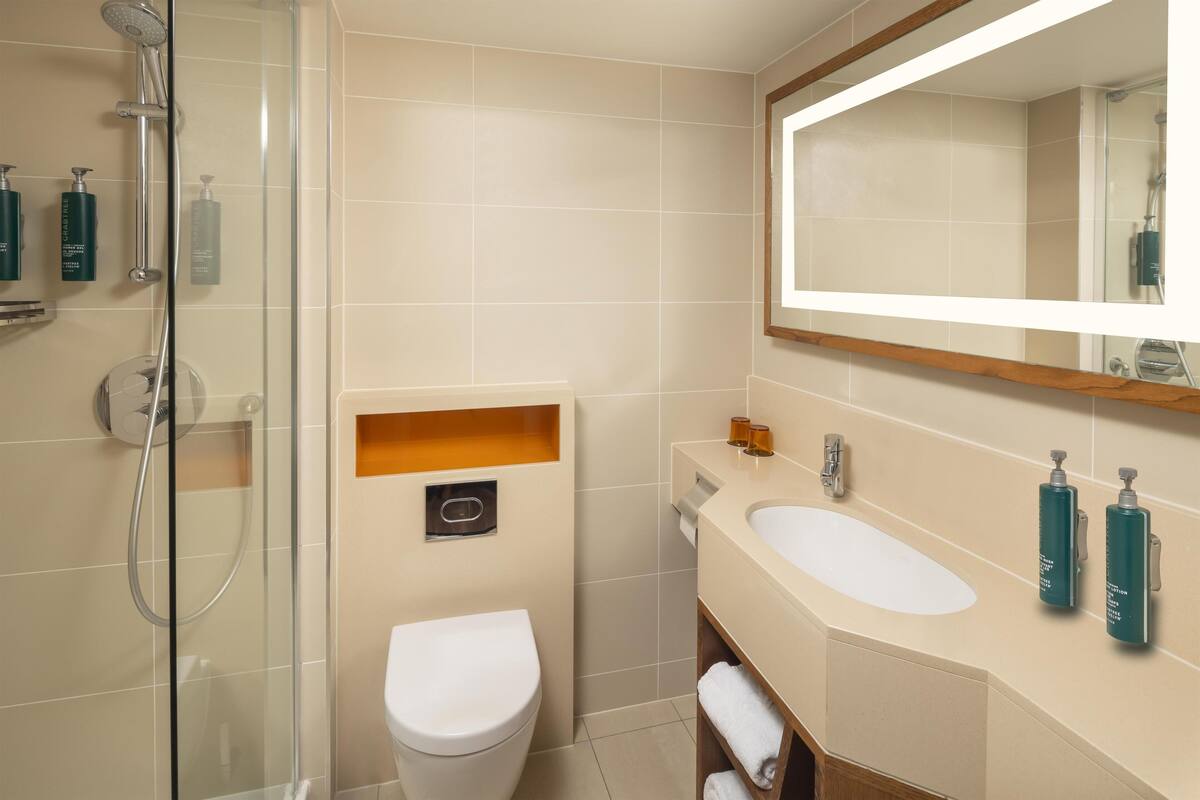 Un baño moderno con cabina de ducha de vidrio, un inodoro blanco, un lavabo color crema con dos botellas de champú y un espejo sobre el lavabo.