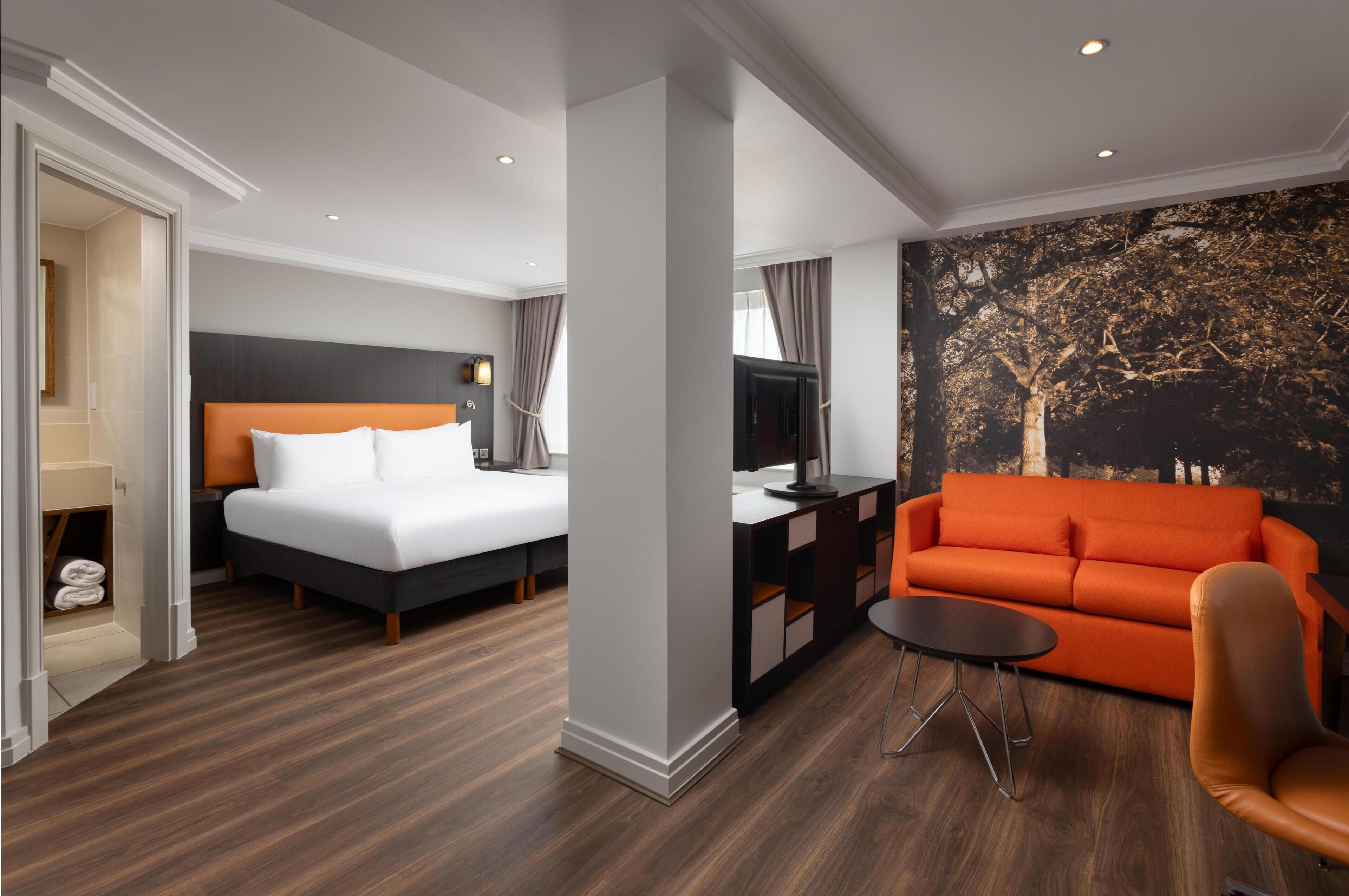 Una habitación de hotel con cama, sofá, una mesita y televisor montado en la pared.