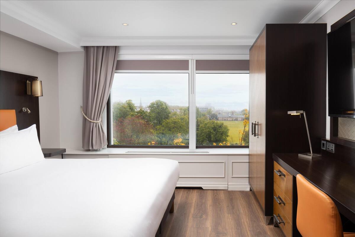 Habitación de hotel con cama, escritorio, televisor y ventana con vista a la ciudad.