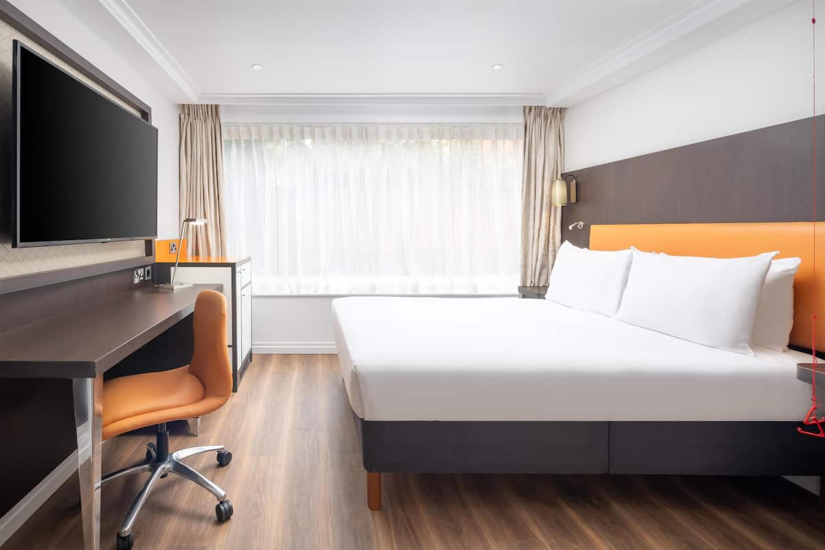 Una habitación de hotel con cama, escritorio con silla, televisor y ventana con cortinas.