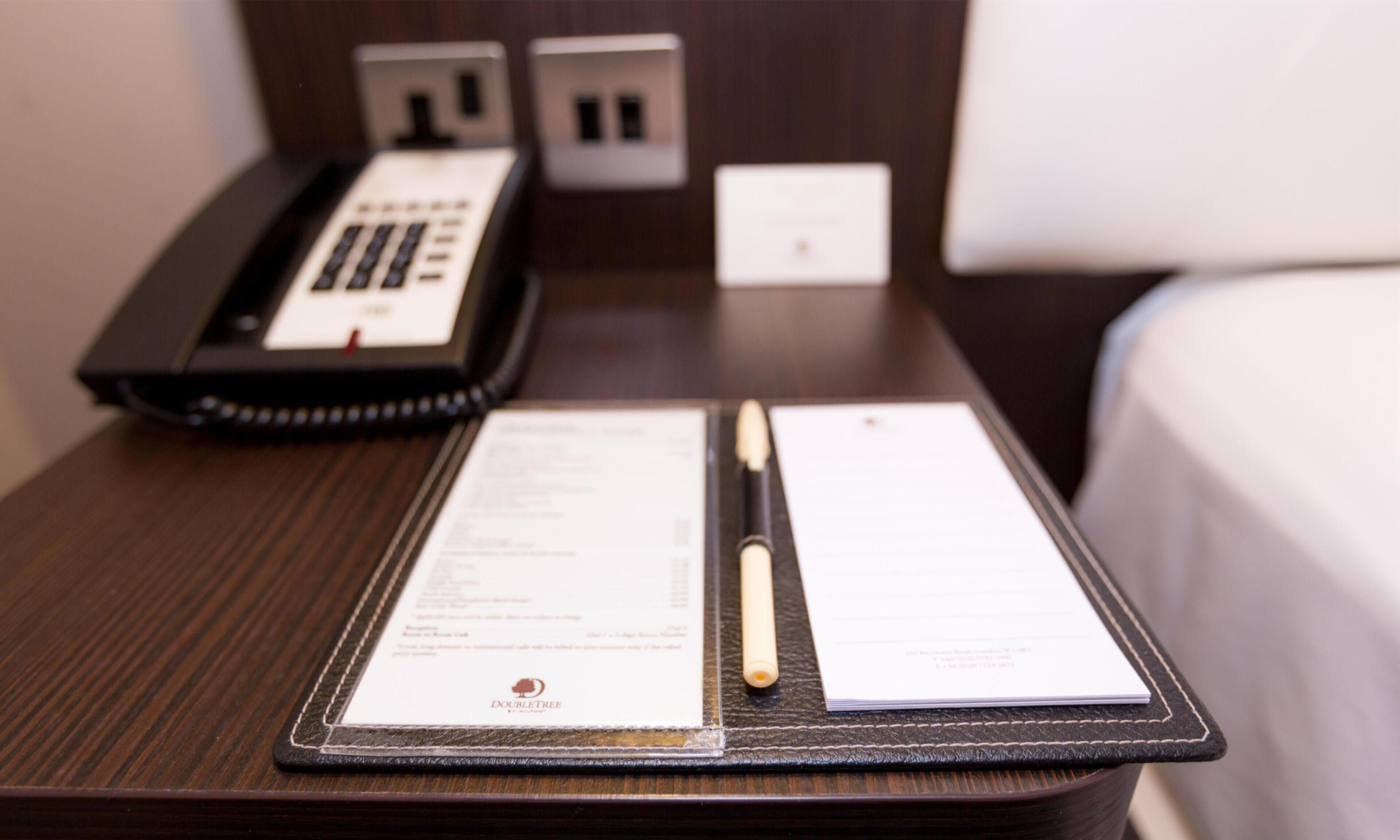 A neatly made bed, a bedside telephone, and a notepad with a pen on a wooden nightstand.