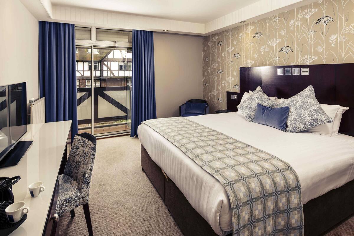 A hotel room with a bed, a desk with a chair, a nightstand, and a view of the outside.