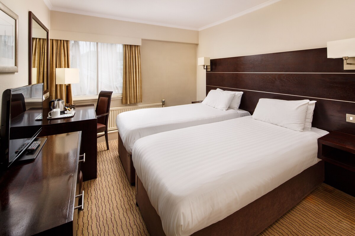 A hotel room with two beds, a desk with a computer, a lamp, and a window with curtains.