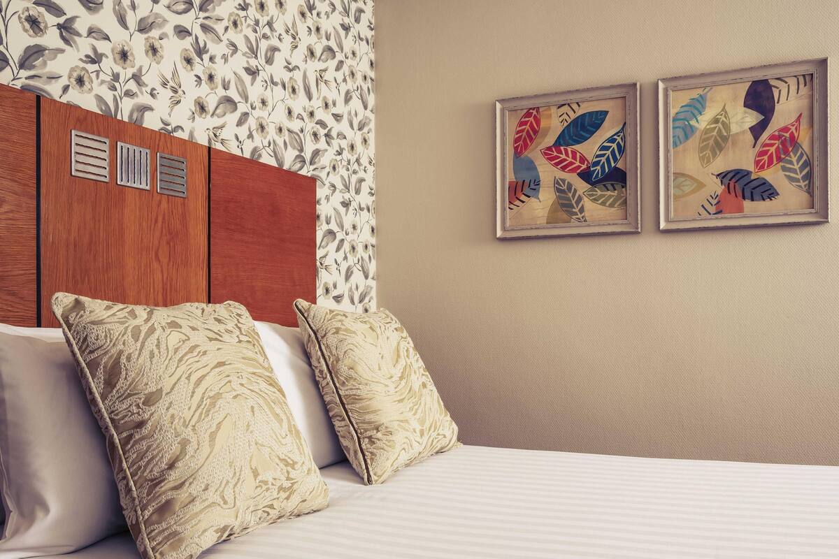 A hotel room with a bed, two decorative pillows, a headboard with ventilation panels, floral wallpaper, and two framed artworks on the wall.