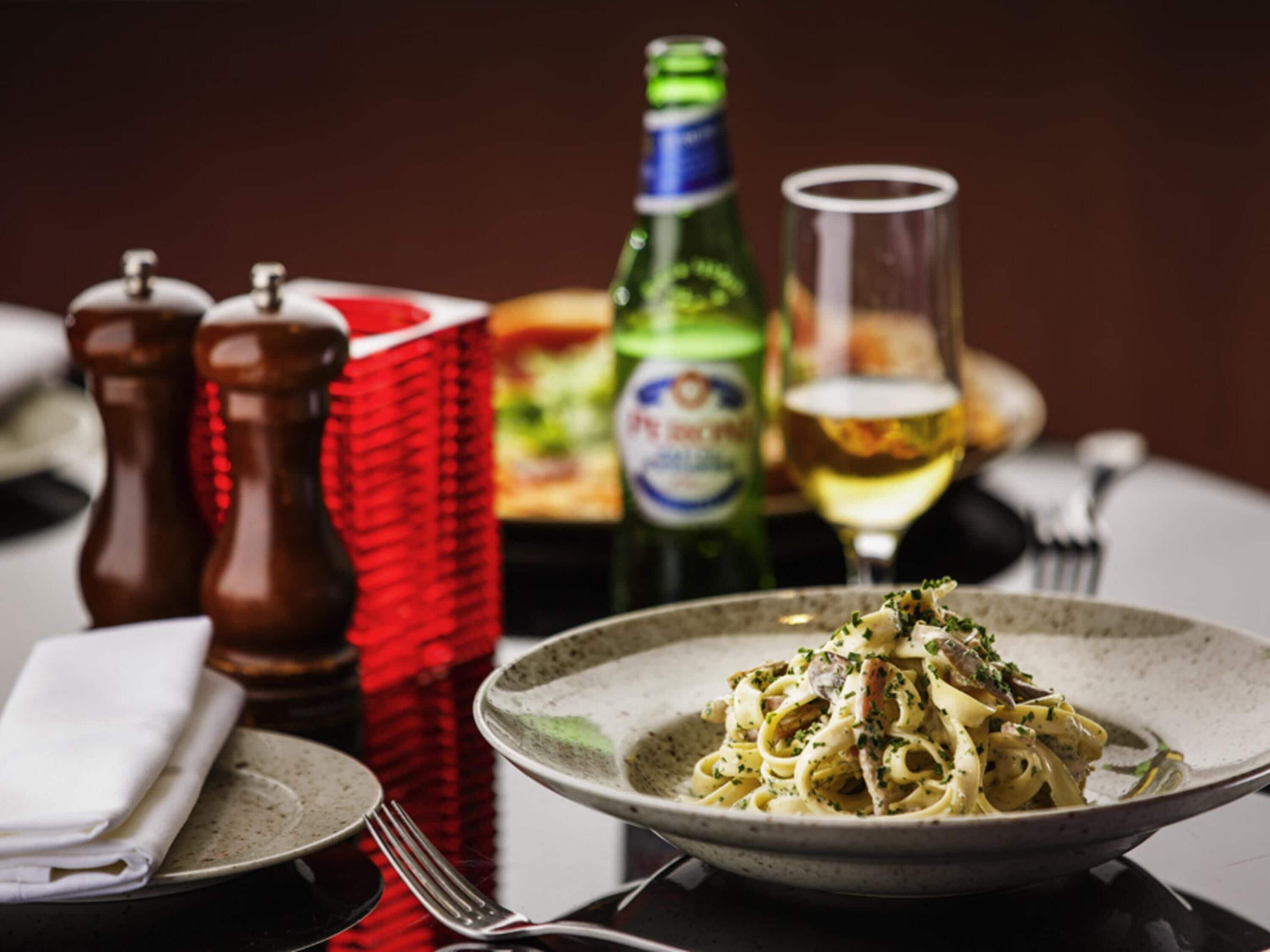 Un plato de pasta con tenedor y cuchillo sobre una mesa, una botella de cerveza y dos saleros.