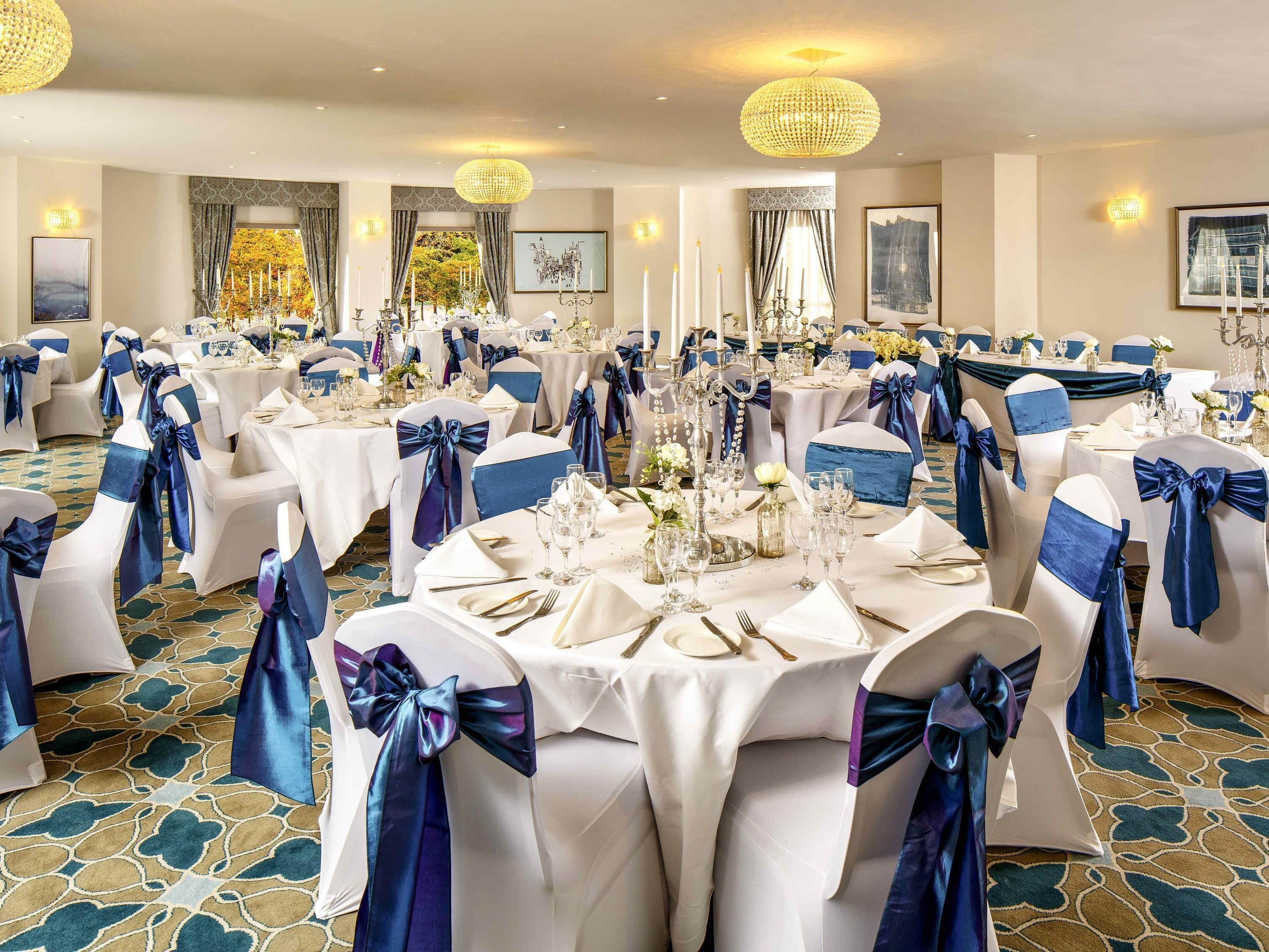 A banquet hall with round tables set for a formal event, featuring white tablecloths and blue sashes on chairs.