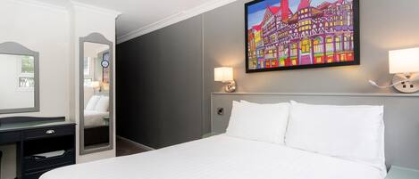 A hotel room with a bed, a desk, a chair, a mirror, a painting of a colorful building, and two bedside lamps.