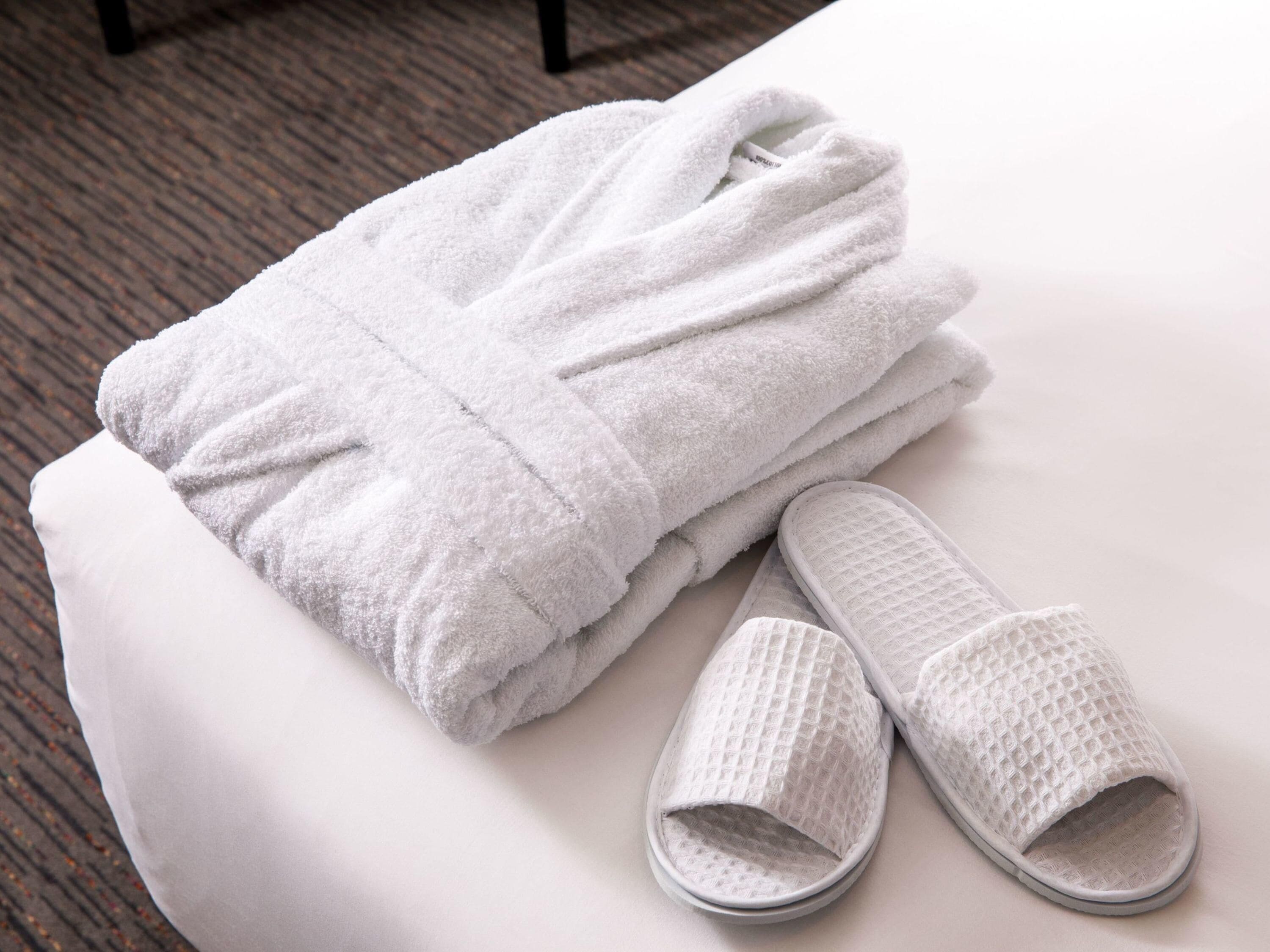 Un albornoz blanco doblado cuidadosamente y un par de pantuflas blancas sobre una superficie blanca.