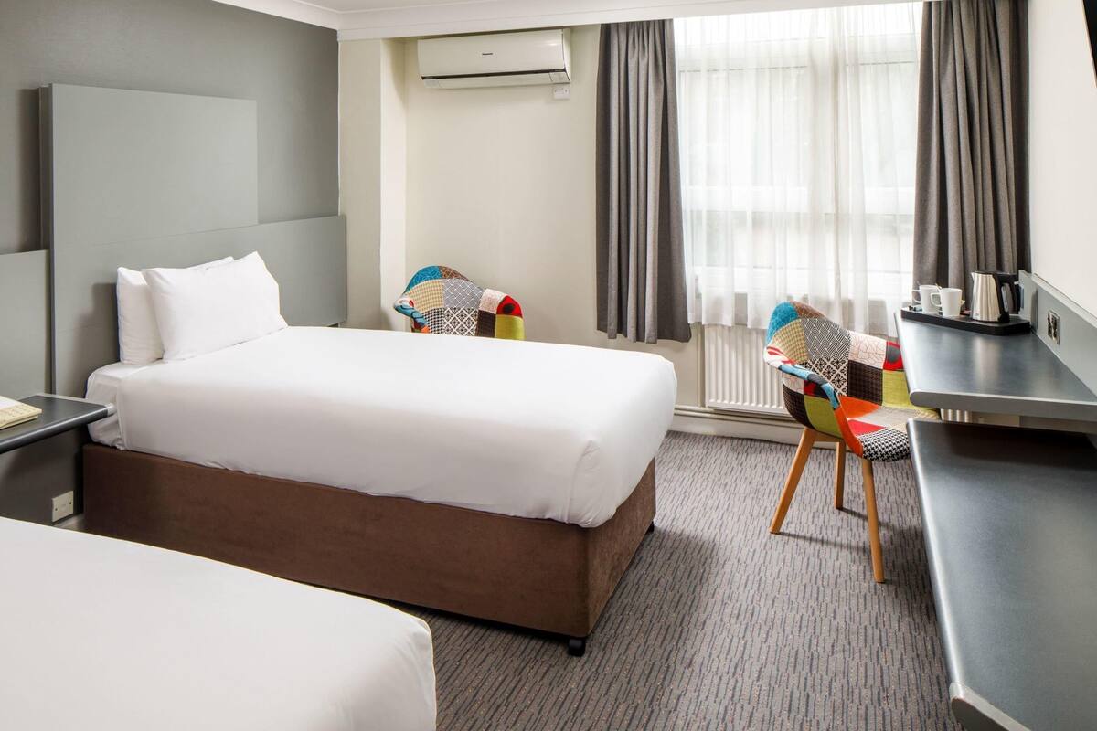 A hotel room with a large bed, a desk with a chair, an air conditioning unit, and a window with curtains.