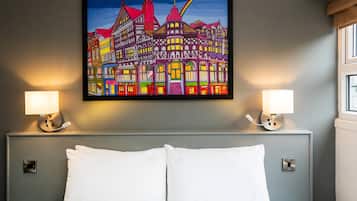 Habitació d'hotel amb un llit, dues làmpades de nit, un quadre penjat a la paret de cases de colors i una finestra amb cortines.