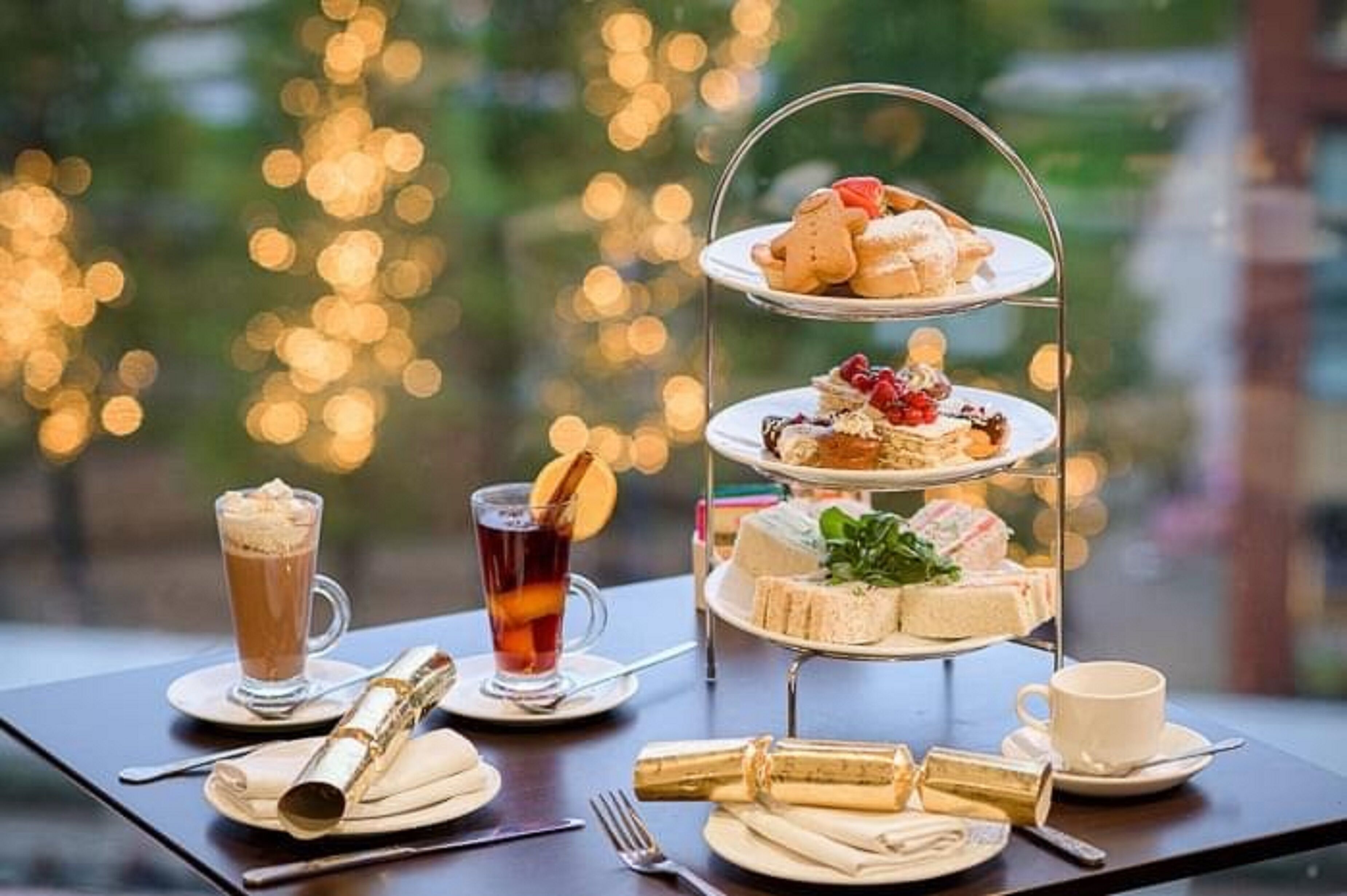 A three-tiered dessert stand with various pastries and sandwiches, a glass of iced tea, a cup of coffee, and a bottle of champagne on a table with a view of a cityscape.