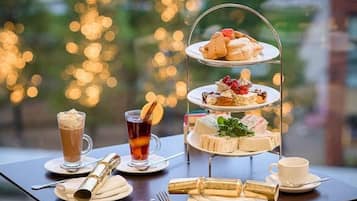 A three-tiered dessert stand with various pastries and sandwiches, a glass of iced tea, a cup of coffee, and a bottle of champagne on a table with a view of a cityscape.