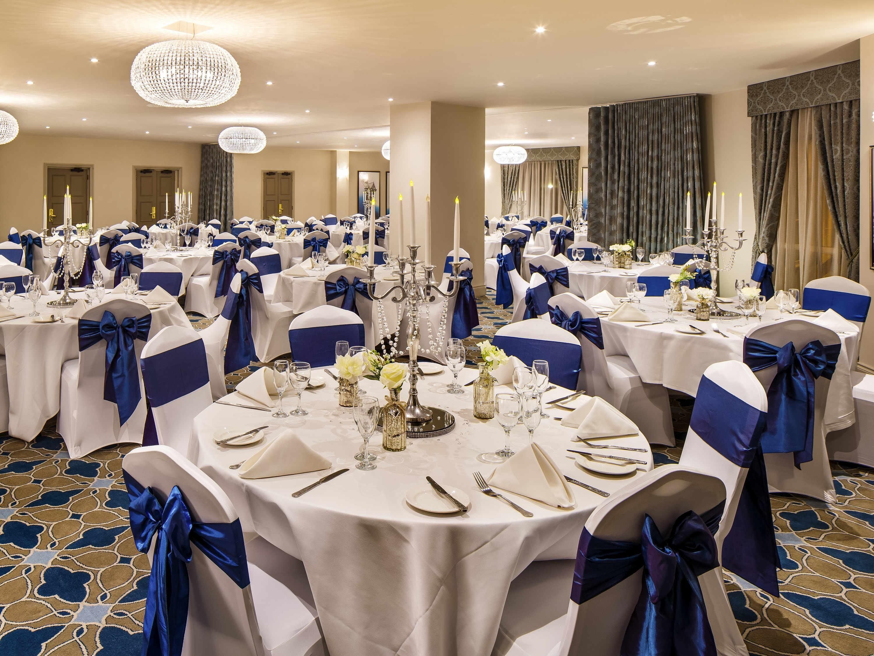 Un salón de banquetes con mesas redondas, cada una con manteles blancos, fajines azules y centros de mesa con velas.