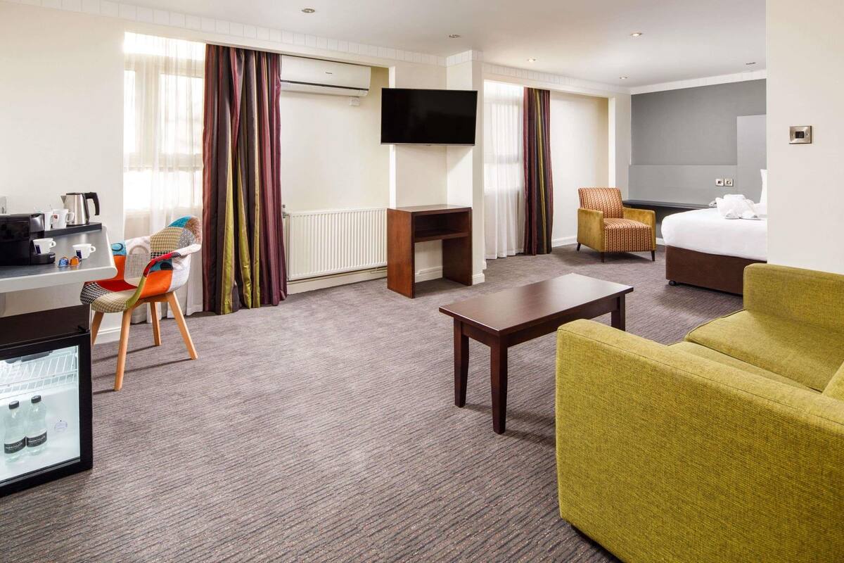 A hotel room with a desk, chair, sofa, TV, bed, and a minibar.