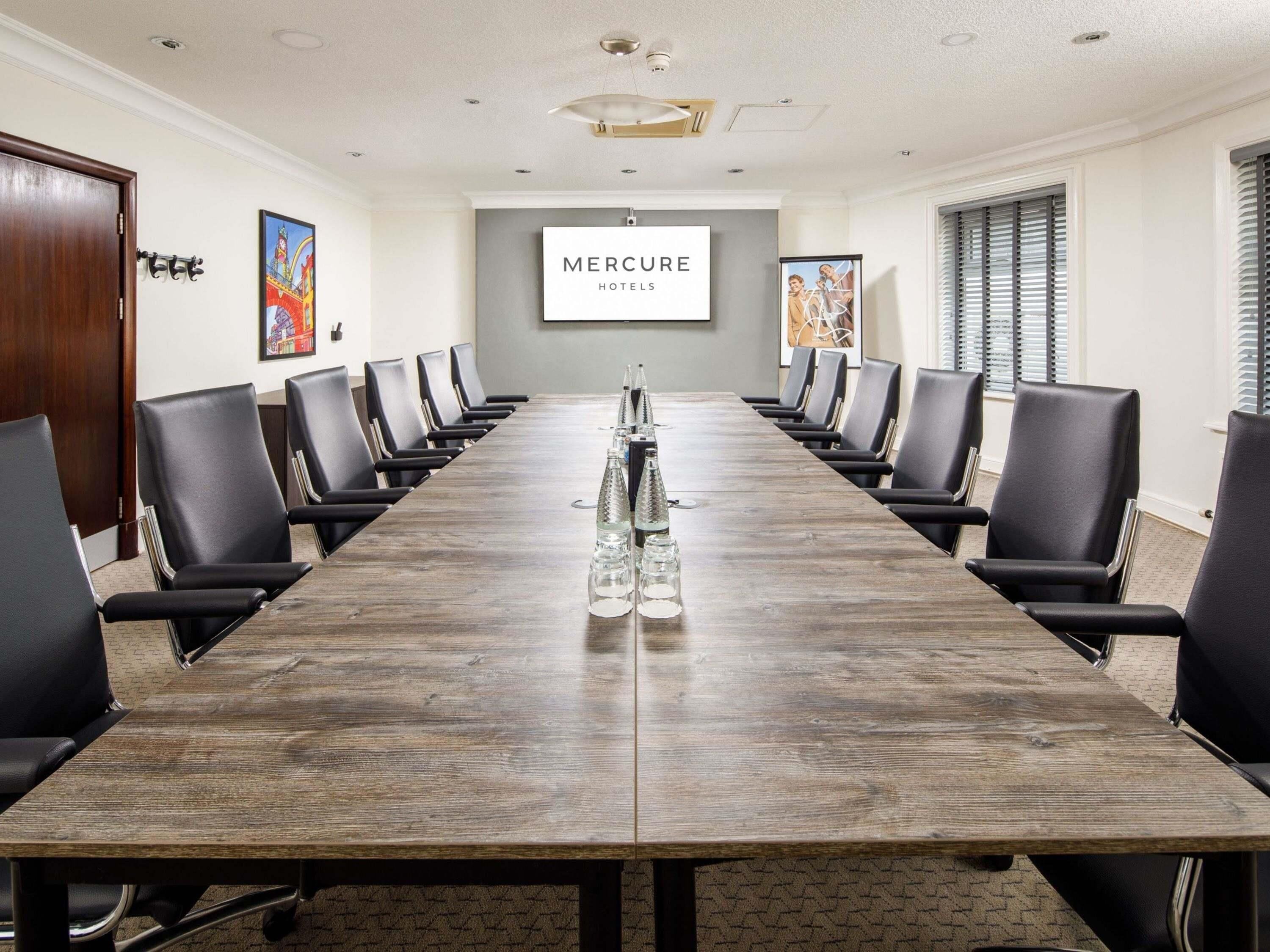 Une salle de conférence avec une longue table, des chaises noires et un écran de projection affichant le logo « Mercure Hotels ».