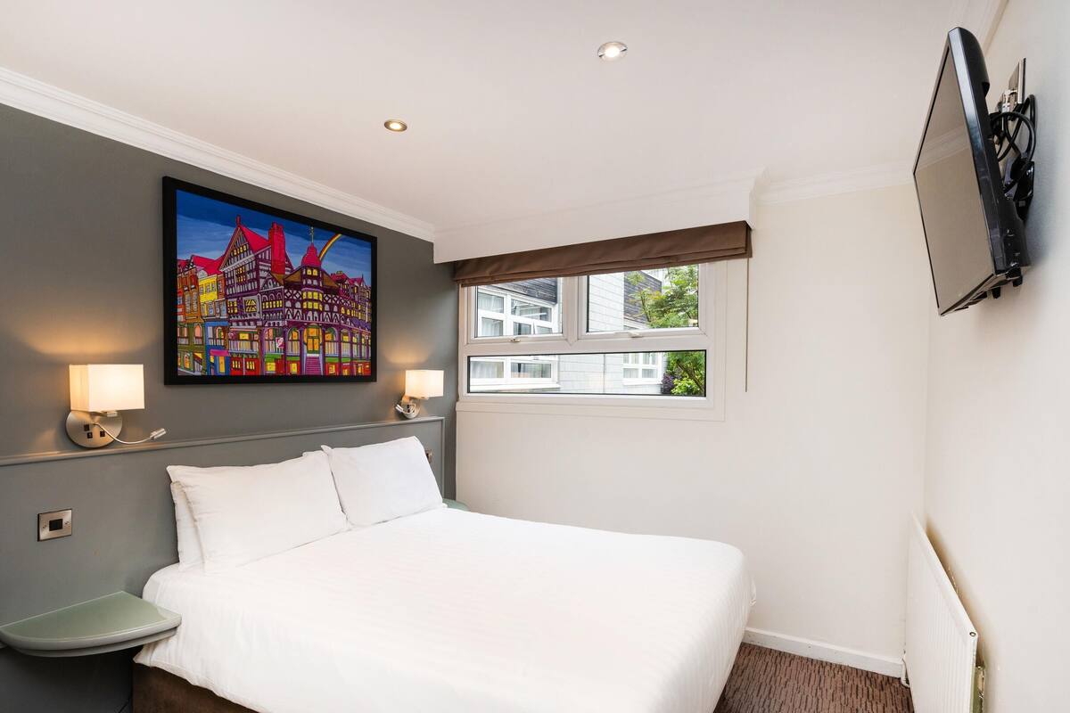 A hotel room with a bed, bedside lamps, a painting, a window, and a TV mounted on the wall.