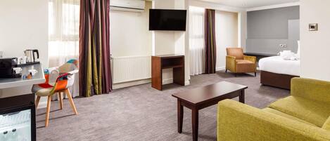 A hotel room with a desk, chair, sofa, TV, bed, and a minibar.